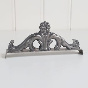 May include: A silver-toned metal napkin holder featuring an arched design with embossed scrollwork and a central flower motif. The base is rectangular with downward-curving ends, providing a stable foundation for holding napkins upright.