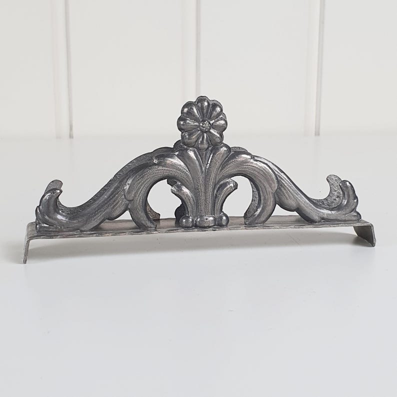 May include: A silver-toned metal napkin holder featuring an arched design with embossed scrollwork and a central flower motif. The base is rectangular with downward-curving ends, providing a stable foundation for holding napkins upright.