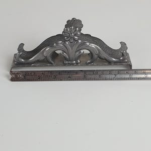 May include: A silver-toned metal napkin holder featuring an ornate floral and scrollwork design. A metal measuring tape is positioned along the base, indicating a length of approximately 6 inches from the left edge to the right side of the holder.