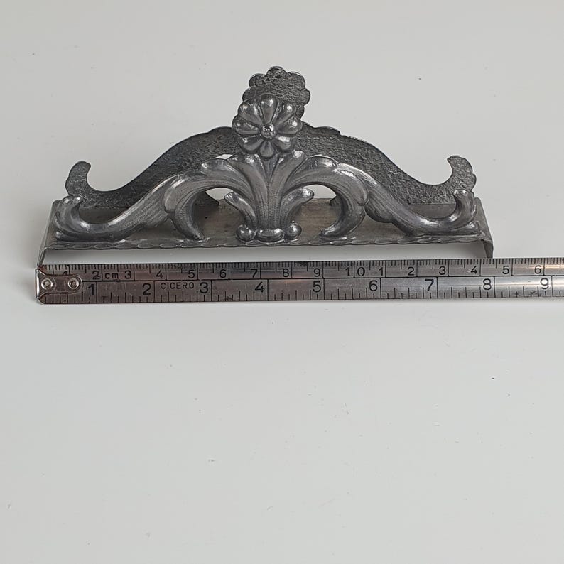 May include: A silver-toned metal napkin holder featuring an ornate floral and scrollwork design. A metal measuring tape is positioned along the base, indicating a length of approximately 6 inches from the left edge to the right side of the holder.