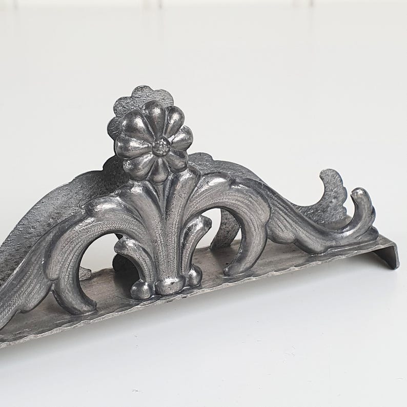 May include: A silver-toned metal napkin holder featuring an ornate, raised floral motif at the center. The design includes symmetrical, curved scrollwork extending outward from the central flower, all set upon a flat, rectangular base.