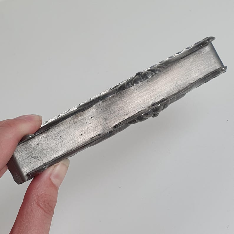 May include: A rectangular, silver-toned metal object with a brushed finish on the side and decorative, rounded, embossed detailing along the edges. The item has a solid, elongated form with a smooth, flat central panel and textured borders.