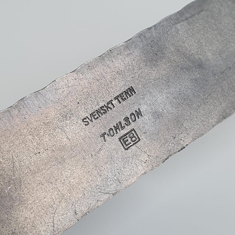 May include: A metallic surface with a hammered, uneven edge featuring stamped text that reads SVENSKT TENN, T. OHLSON, and the letter E inside a square followed by the number 8. The material has a dull, grey, brushed finish with visible scratches.