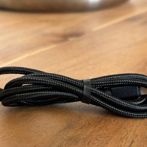 Puede incluir: Cable de carga USB-C negro con diseño trenzado, enrollado sobre una superficie de madera. El cable está asegurado con una banda negra. El cable está diseñado para cargar dispositivos electrónicos.