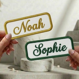 May include: Two embroidered name patches. One features the name "Noah" in gold script on a white background with a gold border. The other has "Sophie" in green script on a white background with a green border. Both are rectangular.