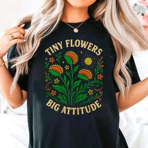 May include: Black t-shirt with a colorful graphic of a Venus flytrap plant. The design includes the text "TINY FLOWERS BIG ATTITUDE" in gold lettering, along with orange, green, and yellow floral elements.