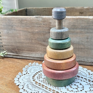 May include: A vintage wooden stacking toy with five rings in shades of green, brown, and pink. The top ring has a gray wooden knob.