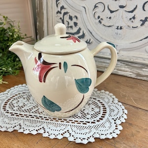 May include: A white ceramic teapot with a floral design featuring pink and blue flowers. The teapot has a handle and a spout. It is sitting on a white lace doily.