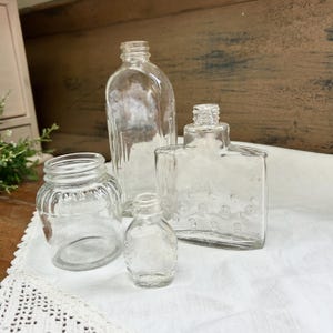 May include: A collection of clear glass bottles and jars of various shapes and sizes. The assortment includes a small, round jar, a tall rectangular bottle, and a square bottle with rounded edges. The objects are arranged on a white surface.