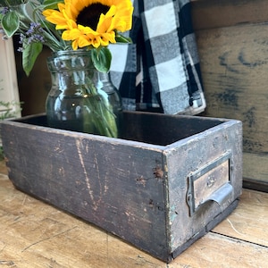 May include: A rectangular, weathered dark brown wooden storage box featuring a metal label holder on the front side. The box contains a clear glass jar filled with bright yellow sunflowers and green foliage, showcasing a rustic, aged wood texture.