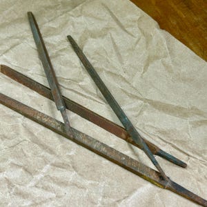 May include: A collection of vintage metal files, showing signs of age and use. The files are various shapes and sizes, with a rusty brown and green patina. They are arranged on a crumpled brown paper surface, suggesting a workshop or tool collection.