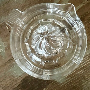 May include: Clear glass citrus juicer with a spiral reamer and a pouring spout. The juicer has a circular design with ridges and a handle. The juicer is made of clear glass.