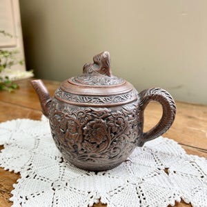 May include: A detailed, bronze-colored teapot with intricate floral and foliate carvings. The teapot features a curved spout, a loop handle, and a decorative lid with a sculpted finial. It rests on a white, crocheted doily, showcasing its ornate design.