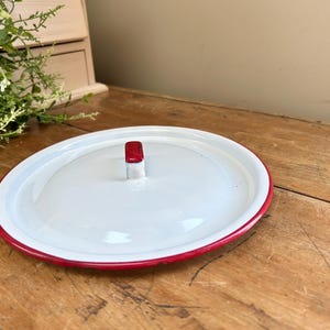 May include: Oval white enamel lid with a red rim and a red handle. The lid is on a wooden surface. The lid is likely for a pot or pan. The lid is a vintage kitchenware item.