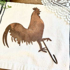May include: A rusty metal rooster silhouette with a hook on the bottom. The rooster is facing right and has a long tail. The hook is perfect for hanging towels or other items.