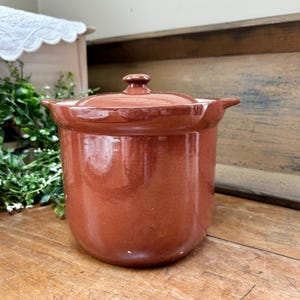 May include: A cylindrical terracotta-colored ceramic pot with a matching lid and two small side handles. The exterior has a smooth, glossy finish with a deep reddish-brown hue. The lid features a small, centered knob handle on top.
