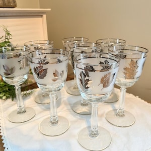 May include: Set of eight clear wine glasses with frosted leaf designs and silver rims. The glasses have long stems and round bases. The leaf designs are in shades of silver and gold. The glasses are arranged on a white cloth.