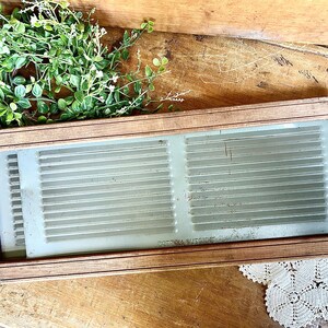 May include: A vintage wooden washboard with a light-colored, ribbed washing surface. The washboard is framed in dark wood and sits on a wooden surface. Greenery is visible in the background.