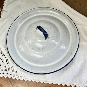 May include: A round, white enamel lid featuring a dark blue rim and a matching dark blue arched handle centered on top. The surface of the lid displays visible signs of wear, including fine scratches and minor surface imperfections throughout.