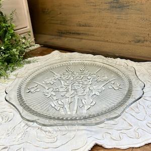 May include: Clear glass cake plate with a floral design. The plate has a scalloped edge and a raised floral pattern in the center. The plate is sitting on a white, textured surface.