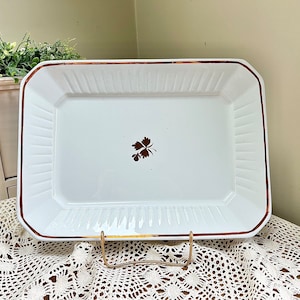 May include: White rectangular baking dish with a gold rim and a brown leaf design in the center. The dish is sitting on a white crocheted tablecloth.