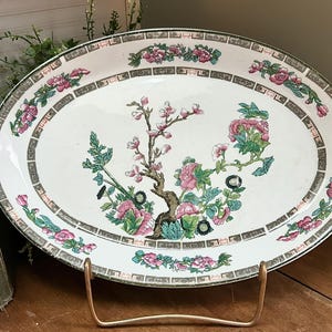 May include: White oval serving platter with a green and pink floral design. The platter has a gold metal stand.