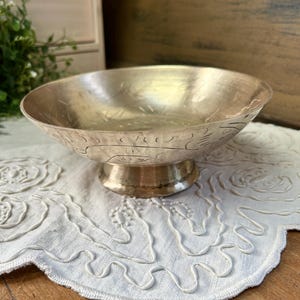 May include: A metallic gold-toned bowl with a pedestal base, featuring etched line patterns along the exterior rim. The bowl has a wide, shallow basin and a smooth, polished finish, resting on a circular footed stand that elevates the main body.
