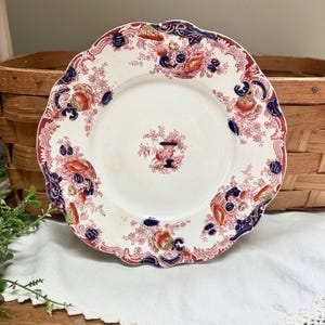 May include: A white antique plate with a scalloped edge, featuring a floral pattern in red, blue, and orange. The plate has a central design and a decorative border. A vintage piece for display or dining.