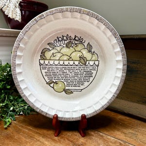 May include: A round ceramic pie dish with a cream-colored base and a brown-striped fluted rim. The center features a green illustration of apples in a bowl with the text "apple pie" and a detailed recipe printed in black ink on the surface.