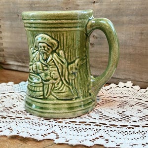 May include: A green ceramic mug with a raised design of a man holding a jug. The mug has a handle and is decorated with green and white glaze.