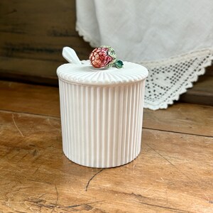 May include: A white ceramic jar with vertical ribbed detailing. The lid is topped with a decorative, colorful raspberry-shaped knob. A small white spoon rests on the lid. The jar is on a wooden surface.