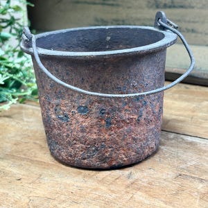 May include: A small, rustic metal bucket with a handle. The bucket has a weathered, textured appearance with shades of brown and rust. The handle is made of a thin, silver-colored metal. The bucket sits on a wooden surface.