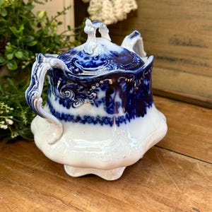 May include: A vintage white and blue ceramic sugar bowl with a lid. The bowl features a detailed floral design with gold accents. The handles and lid finial are intricately shaped. The bowl is on a wooden surface.