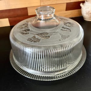 May include: A clear glass cake stand with a domed lid. The lid features an embossed floral design and a textured knob. The base has a ribbed pattern and a decorative edge, perfect for displaying baked goods.