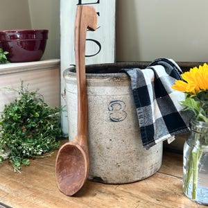 May include: A long, hand-carved wooden spoon with a curved handle. The spoon is a warm brown color and has a rustic, farmhouse aesthetic. It is displayed next to a vintage stoneware crock with the number "3" on it.