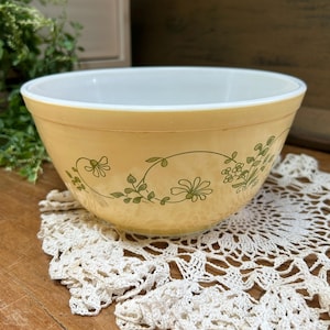 May include: A round mixing bowl with a pale yellow exterior and a white interior. The exterior features a repeating green line art pattern of delicate flowers, stems, and leaves encircling the bowl. The bowl has a smooth, glossy finish.