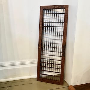 May include: A rectangular metal grate featuring a grid pattern with a weathered, rusted brown finish. The frame is solid and surrounds a series of intersecting horizontal and vertical bars, creating a uniform square mesh design throughout the piece.