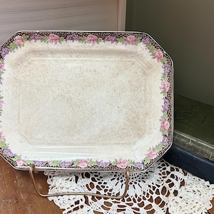 May include: A white ceramic serving platter with a floral design in shades of pink, purple, and brown. The platter has a scalloped edge and is on a metal stand.