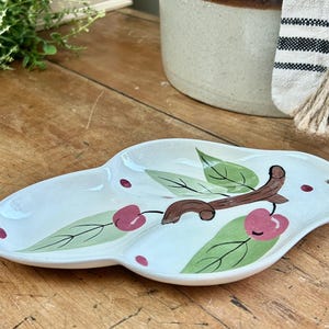 May include: A white ceramic serving dish with a cherry branch design. The dish has three sections and is decorated with green leaves and red cherries.