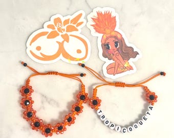 May include: Two orange flower bracelets with black centers and adjustable cords. One bracelet spells out "TROPICOQUETA" in white letter beads. Two orange stickers with illustrations of a flower and a cartoon woman are also present.