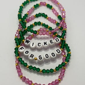 May include: Stack of beaded bracelets in shades of pink and green with gold-colored accents. The bracelets spell out "WICKED FOR GOOD" in white letter beads. The bracelets are on a white background.