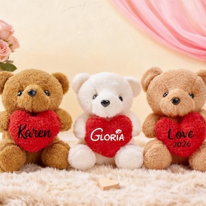 Pode incluir: Três ursos de pelúcia com corações vermelhos. Um urso marrom com "Karen", um branco com "Gloria" e outro marrom com "Love 2026". Estão sobre uma superfície macia. Rosas num vaso são visíveis no fundo.