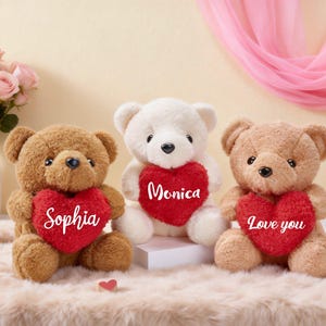 May include: Three teddy bears in shades of brown and white, each clutching a red heart. The hearts are inscribed with "Sophia", "Monica", and "Love you" in white script. A vase of pink roses and a small angel statue are in the background.