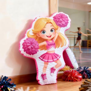 Può includere: Un cuscino in peluche sagomato a forma di cheerleader dei cartoni animati con capelli biondi, che indossa un'uniforme rosa e bianca con il nome Basia stampato sul petto. Il personaggio tiene due pon pon rosa e indossa scarpe da ginnastica.