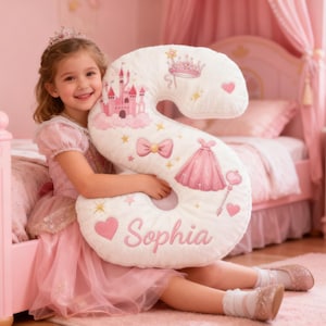 May include: A white, plush letter "S" cushion decorated with pink castle, dress, and crown motifs. The cushion is embroidered with the name "Sophia" in pink. The girl is wearing a pink dress and tiara, holding the cushion in a pink bedroom.