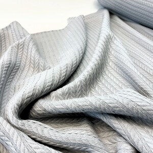 May include: Close-up of a light gray fabric with a cable knit pattern. The fabric is draped, showing the texture and weave of the material. A roll of the same fabric is visible in the background.