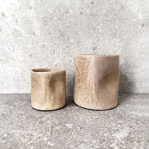 Può includere: Due tazze cilindriche in ceramica con una finitura testurizzata e maculata nei toni del beige e del marrone chiaro. La tazza più piccola è posizionata a sinistra, mentre quella più grande si trova a destra. Entrambi i recipienti hanno una forma organica.