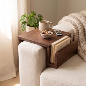 May include: A dark brown wooden sofa table with a built-in book holder. The table holds a beige mug, a small bowl of nuts, and a remote control. Two books are in the book holder. A green plant and a blanket are in the background.