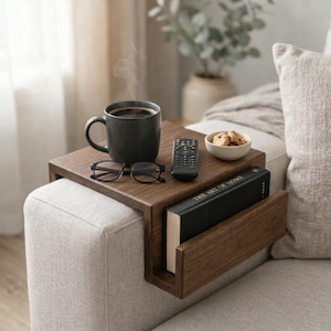 Puede incluir: Una bandeja de brazo de sofá de madera oscura con un sujetalibros incorporado. Una taza de café, gafas, un mando a distancia y un cuenco pequeño con galletas están en la bandeja. Un libro titulado "THE ART OF HOME" está en el soporte.