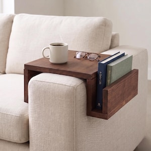 May include: A dark brown wooden sofa arm table with a built-in book holder. A white mug with coffee and a pair of glasses rest on the table. Two books are in the holder. The table is placed on the arm of a beige sofa.